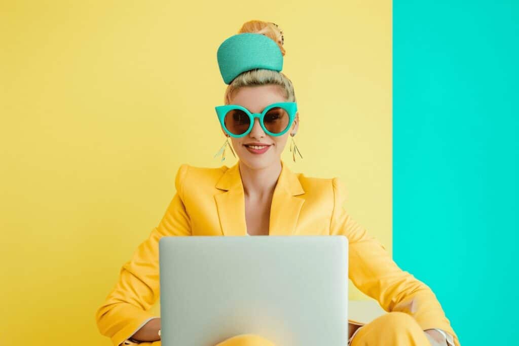 A woman in a yellow suit with sunglasses and a laptop on a yellow and blue background.