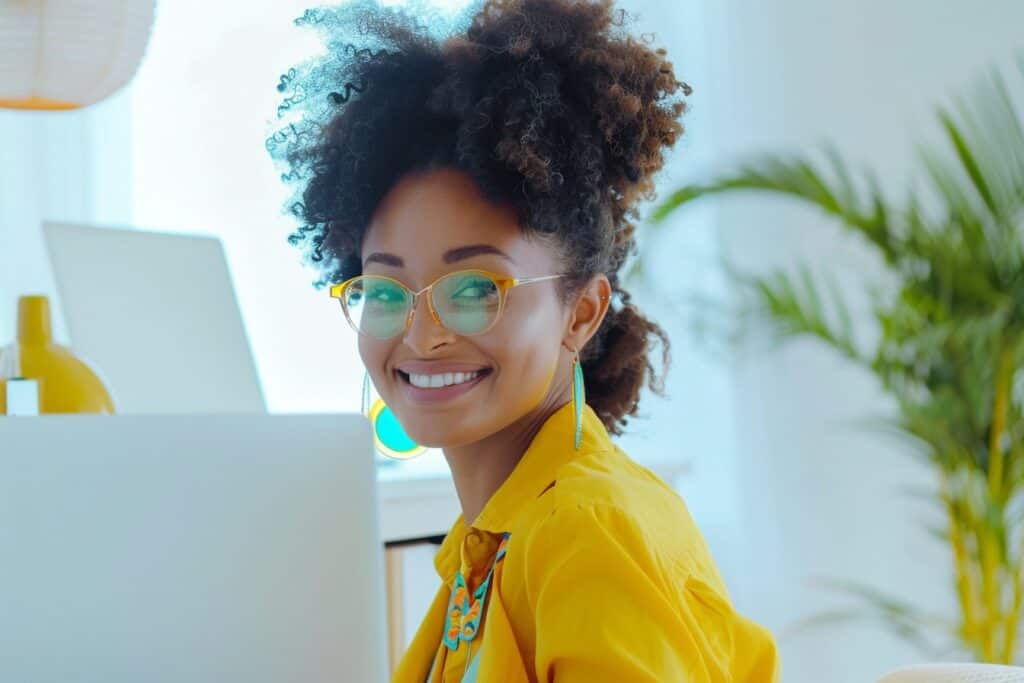 A person with curly hair, wearing glasses and a yellow shirt, smiling while sitting at a desk with a laptop engaged in AI content creation. There is a green plant in the background.
